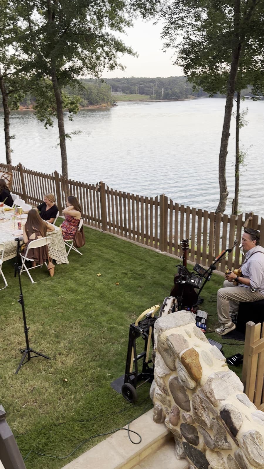Guests seated at long tables beside the lake at dusk while a musician plays